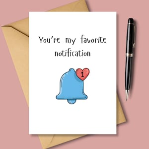 Puede incluir: Tarjeta de felicitación blanca con el texto "You're my favorite notification" y un icono de campana azul con un corazón rojo y el número 1. Un bolígrafo negro y un sobre marrón también son visibles.