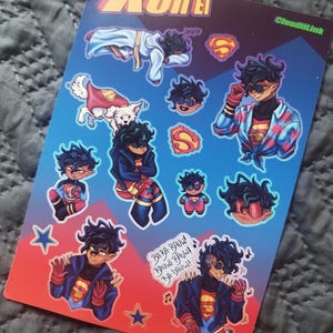 May include: A sticker sheet featuring multiple illustrations of Kon El, a character with dark hair and sunglasses, in various poses. The sheet has a blue and red gradient background with the text "Kon El" at the top. Includes the text "BA-BA-BAUW! BAUWI BAUW! BA-DAUW!"