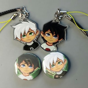 Pode incluir: Dois pingentes de acrílico transparente e dois botões redondos com personagens de desenhos animados com cabelo branco e preto, olhos verdes e azuis e camisetas. Os pingentes têm cordões pretos e fechos prateados. Os botões têm fundo verde.