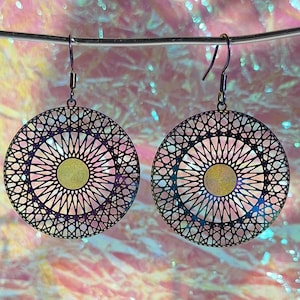 Könnte beinhalten: Ein Paar kreisförmige Ohrhänger mit einem dunklen, irisierenden Metall-Filigranmuster in einem strahlenden geometrischen Design. Jeder Ohrring hat eine massive, kreisförmige goldfarbene Mitte und ist an einem einfachen Metallhaken befestigt.
