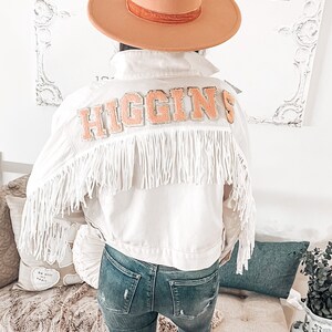 Peut inclure: Veste en jean blanche avec des franges et un patch "HIGGINS" de couleur p&ecirc;che au dos. La veste est port&eacute;e sur un jean bleu.