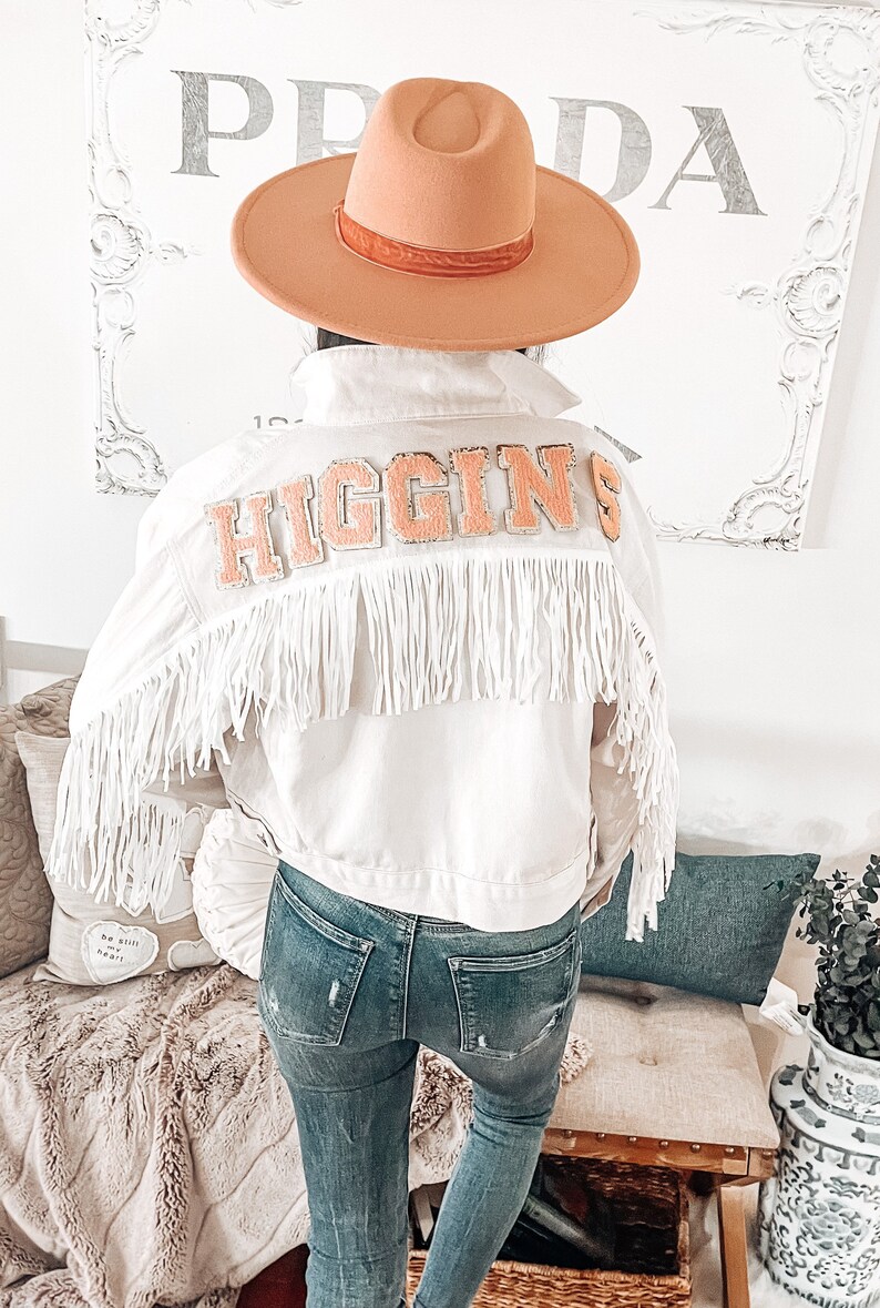 Peut inclure: Veste en jean blanche avec des franges et un patch "HIGGINS" de couleur p&ecirc;che au dos. La veste est port&eacute;e sur un jean bleu.