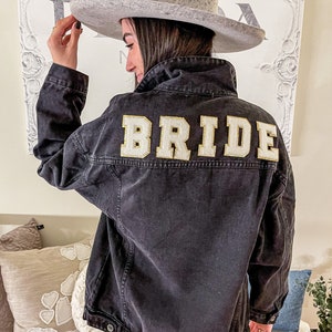 Pu&ograve; includere: Una giacca di jeans nera con la parola "BRIDE" ricamata sul retro in lettere dorate. La giacca &egrave; indossata da una persona che indossa un cappello grigio e jeans blu.
