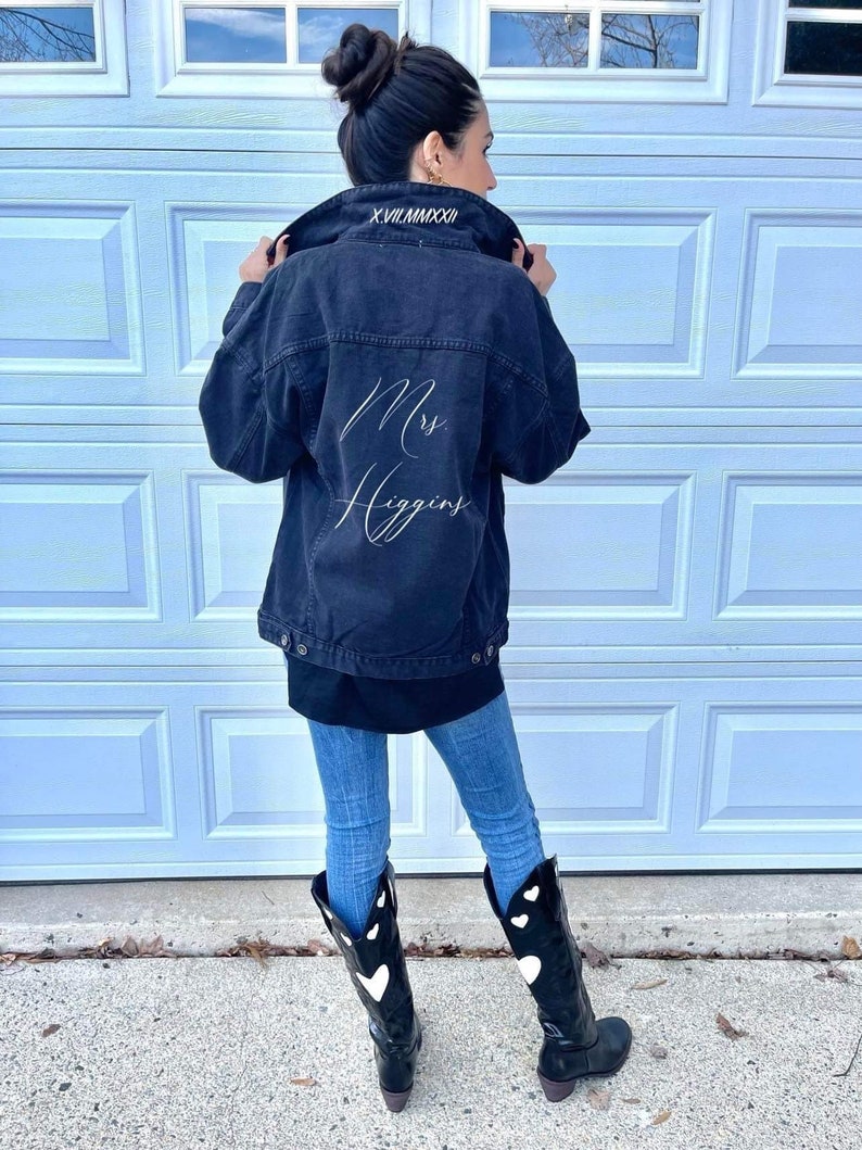 Pu&ograve; includere: Una giacca di jeans nera con ricamo bianco che dice "Mrs. Higgins" sul retro. La giacca ha un colletto nero con cuciture bianche che dicono "X.VII.MMXXVII". La persona che indossa la giacca indossa anche jeans blu e stivali neri con cuori bianchi.