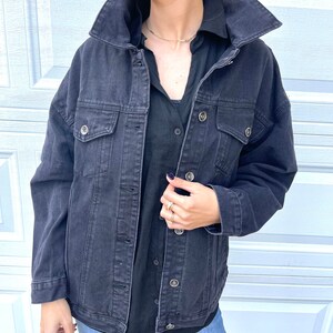 Peut inclure: Une veste en jean noire avec un col, des manches longues et plusieurs poches. La veste est d&eacute;boutonn&eacute;e et la personne qui la porte tient le devant de la veste.