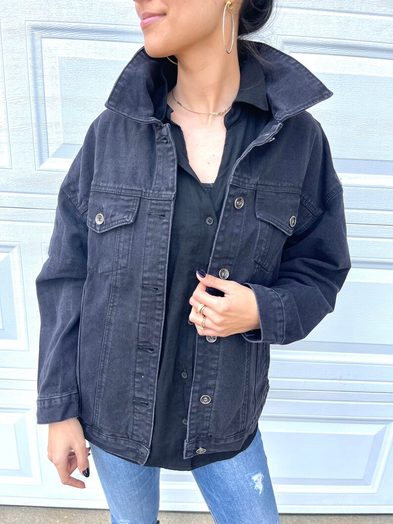 Peut inclure: Une veste en jean noire avec un col, des manches longues et plusieurs poches. La veste est d&eacute;boutonn&eacute;e et la personne qui la porte tient le devant de la veste.