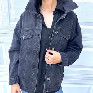 Pu&ograve; includere: Una giacca di jeans nera con colletto, maniche lunghe e diverse tasche. La giacca &egrave; sbottonata e la persona che la indossa tiene la parte anteriore della giacca.