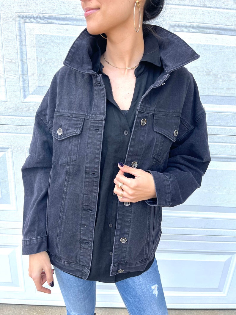 Pu&ograve; includere: Una giacca di jeans nera con colletto, maniche lunghe e diverse tasche. La giacca &egrave; sbottonata e la persona che la indossa tiene la parte anteriore della giacca.