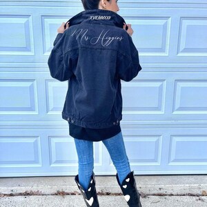Pu&ograve; includere: Una giacca di jeans nera con ricamo bianco che dice "Mrs. Higgins" sul retro. La giacca ha un colletto nero con ricamo bianco che dice "XVII.MMXXI".