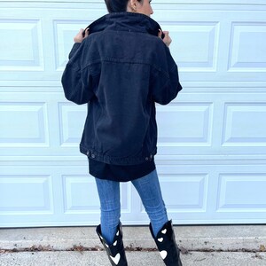 Peut inclure: Une veste en jean noire &agrave; la coupe d&eacute;contract&eacute;e port&eacute;e sur un t-shirt noir &agrave; manches longues. La personne porte un jean bleu et des bottes de cow-boy noires avec des motifs de c&oelig;urs blancs.
