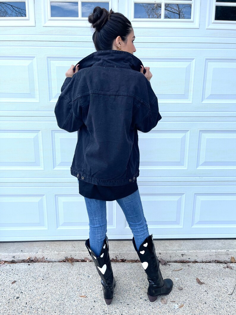 Peut inclure: Une veste en jean noire &agrave; la coupe d&eacute;contract&eacute;e port&eacute;e sur un t-shirt noir &agrave; manches longues. La personne porte un jean bleu et des bottes de cow-boy noires avec des motifs de c&oelig;urs blancs.