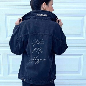 Pu&ograve; includere: Una giacca di jeans nera con ricamo bianco sul retro che dice "Future Mrs. Higgins" e sul colletto dice "XVII.VIII.MMXXII".