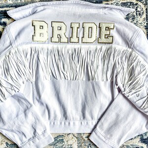Peut inclure: Veste en jean blanche avec des franges et un patch "BRIDE" en paillettes dor&eacute;es au dos.