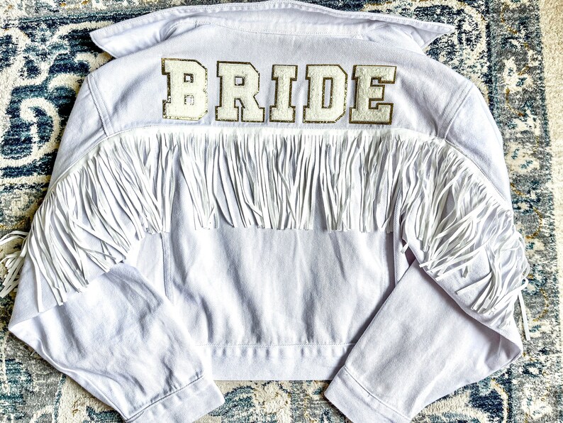 Peut inclure: Veste en jean blanche avec des franges et un patch "BRIDE" en paillettes dor&eacute;es au dos.