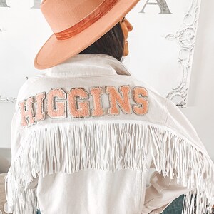 Peut inclure: Une veste en jean blanche avec des d&eacute;tails en franges et un patch en feutre rose qui dit "HIGGINS" au dos. La veste est port&eacute;e sur un jean bleu et une personne porte un chapeau &agrave; large bord.