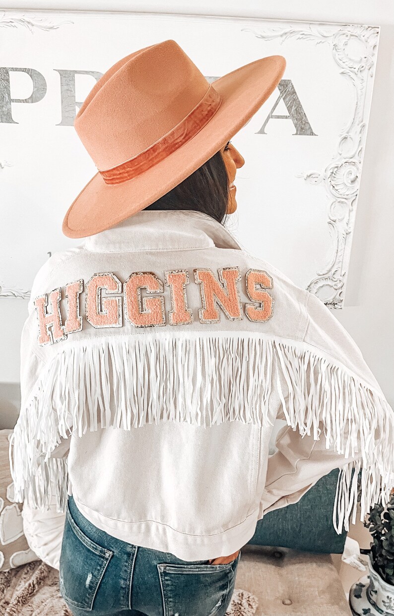 Peut inclure: Une veste en jean blanche avec des d&eacute;tails en franges et un patch en feutre rose qui dit "HIGGINS" au dos. La veste est port&eacute;e sur un jean bleu et une personne porte un chapeau &agrave; large bord.
