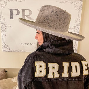 Peut inclure: Une femme portant une veste en jean noire avec le mot "BRIDE" brod&eacute; au dos en lettres blanches avec des bordures dor&eacute;es. Elle porte un chapeau fedora en feutre gris.