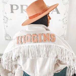 Peut inclure: Une veste en jean blanche avec une bordure en franges roses et un patch rose "HIGGINS" au dos. La veste est port&eacute;e sur un jean bleu et une personne porte un chapeau &agrave; large bord.