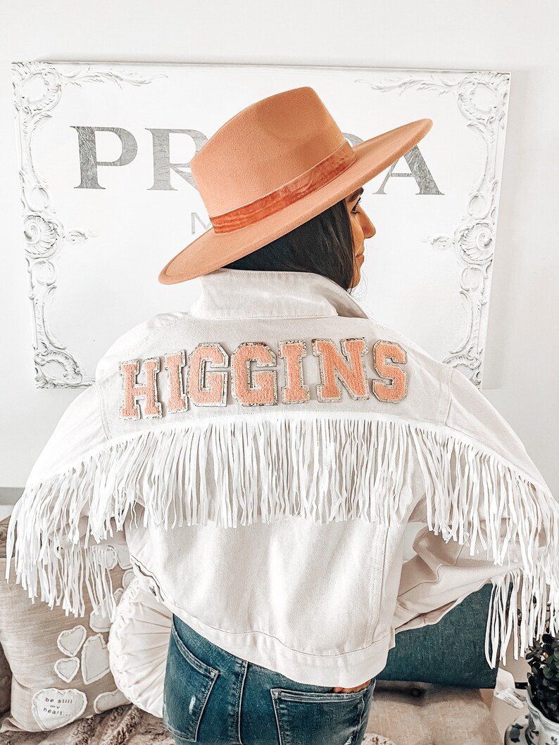 Peut inclure: Une veste en jean blanche avec une bordure en franges roses et un patch rose "HIGGINS" au dos. La veste est port&eacute;e sur un jean bleu et une personne porte un chapeau &agrave; large bord.
