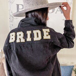 Peut inclure: Une veste en jean noire avec le mot "BRIDE" brod&eacute; en or au dos. La veste est port&eacute;e par une personne portant un fedora gris.