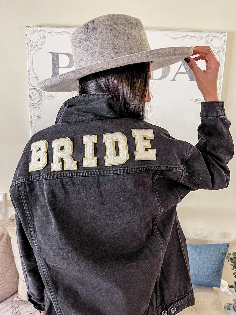 Peut inclure: Une veste en jean noire avec le mot "BRIDE" brod&eacute; en or au dos. La veste est port&eacute;e par une personne portant un fedora gris.