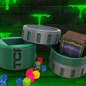 May include: Green and gray gaming accessories, including dice and card holders, are displayed against a green and black brick wall background. The card holder contains a deck of cards. Colorful dice are scattered around the holders.