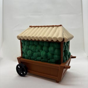 Op de afbeelding: Een miniatuur bruine kar met een beige dak en een zwart wiel. De kar is gevuld met groene bollen. Het dak heeft een geschulpte rand. De kar heeft een traditioneel Aziatisch ontwerp.