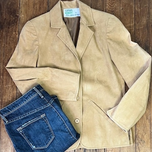 May include: A tan suede blazer featuring a notched lapel, two front buttons, and two welt pockets. The interior label reads Samuel Robert by Pepi's Made in USA. Paired with dark blue denim jeans featuring a classic five-pocket design and stitching.