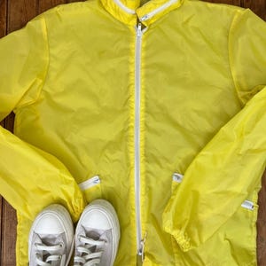 May include: A bright yellow windbreaker jacket with a white front zipper, a high collar, and two zippered side pockets. The interior neck label reads Macy's men's store. A pair of light gray canvas sneakers with white laces and rubber soles are positioned below.