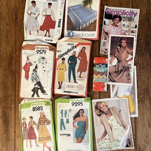 May include: A collection of vintage sewing patterns and magazines, including McCall's and Simplicity brands. The patterns feature illustrations of clothing designs, and the magazines showcase fashion trends. The items are arranged on a wooden surface.