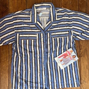 May include: A short-sleeved button-down shirt featuring a pattern of vertical blue, white, and black stripes with small brown and blue chevron accents. The shirt includes a collar, two chest pockets with flaps, and a tag showing a clothing illustration.