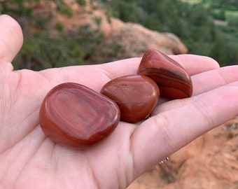 May include: Three red jasper tumbled stones with brown and white stripes. The stones are in a person's hand.