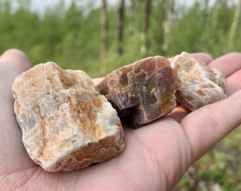 May include: Three rough, natural, brown and white crystals in a hand. The crystals are translucent and have a rough, uneven texture.