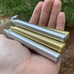 Könnte beinhalten: Drei Wachsstangen, eine silberne, eine goldene und eine silberne, liegen übereinander. Die Stangen sind etwa 15 cm lang und haben eine glatte, glänzende Oberfläche.