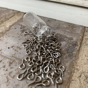 Könnte beinhalten: Ein kleines Glasgefäß verschüttet eine große Menge silberfarbener Ösenhaken. Die Haken liegen verstreut auf einer strukturierten braunen Oberfläche.