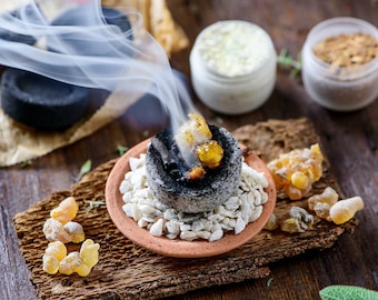 May include: A close-up of a burning incense with white smoke rising from a small clay dish. The dish is filled with white sand and a black charcoal disc. There are small pieces of amber resin scattered around the dish.