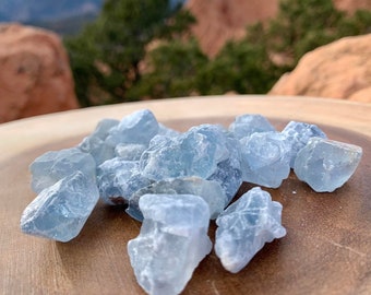 May include: A collection of light blue celestite crystals on a wooden surface. The crystals are rough and have a natural, unpolished look.