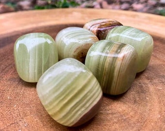 May include: A group of six green and brown tumbled stones on a wooden surface. The stones are smooth and polished, with a variety of patterns and textures.