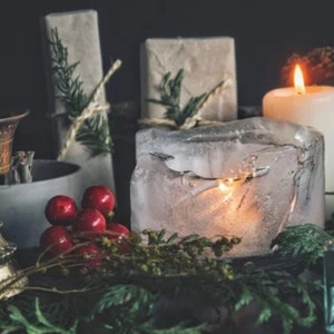May include: A lit candle sits inside a block of ice, surrounded by greenery and red berries. The candle is white and the ice is clear. There are two wrapped packages in the background.
