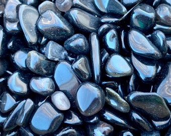 May include: A close-up of a pile of black obsidian tumbled stones. The stones are smooth and polished, with a deep black color and a slight sheen.