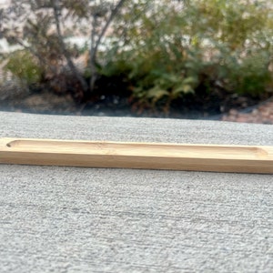 May include: A natural wood incense holder with a rectangular shape and a slightly rounded edge. The holder is made of light brown wood and is sitting on a gray concrete surface.