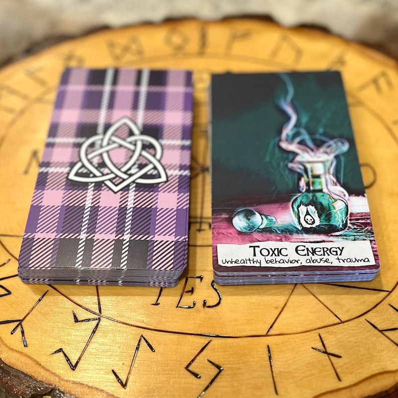 May include: Two tarot cards on a wooden surface. The card on the left has a pink and purple plaid pattern with a white Celtic knot design. The card on the right has a dark green background with a glass bottle with a skull and crossbones design. The text on the card reads "Toxic Energy, unhealthy behavior, abuse, trauma".
