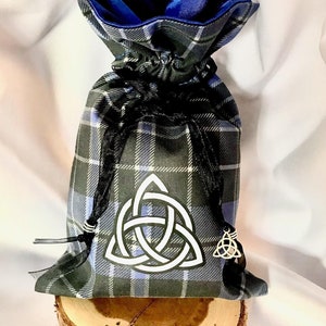 May include: A blue and grey plaid drawstring bag with a white Celtic knot design and a blue satin lining. The bag is sitting on a wooden slice.