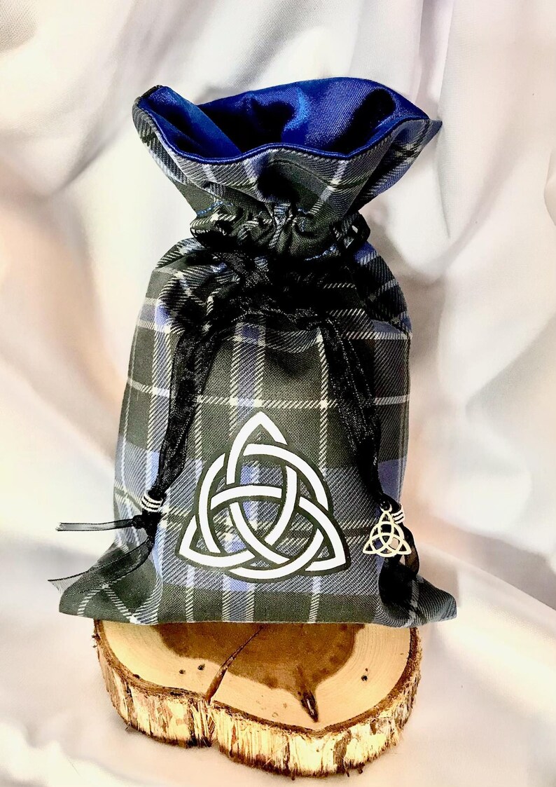 May include: A blue and grey plaid drawstring bag with a white Celtic knot design and a blue satin lining. The bag is sitting on a wooden slice.