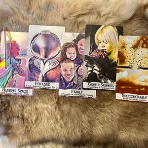 May include: Five tarot cards with colorful illustrations and handwritten text. The cards are laid out on a brown fuzzy surface. The cards are titled "Focused", "Grief & Sadness", "Needing Space", "Family", and "Irreconcilable".