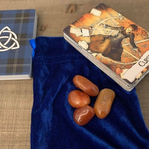 May include: Four carnelian stones, a type of quartz, are arranged on a blue velvet pouch. The stones are a reddish-brown colour and have a smooth, polished finish.