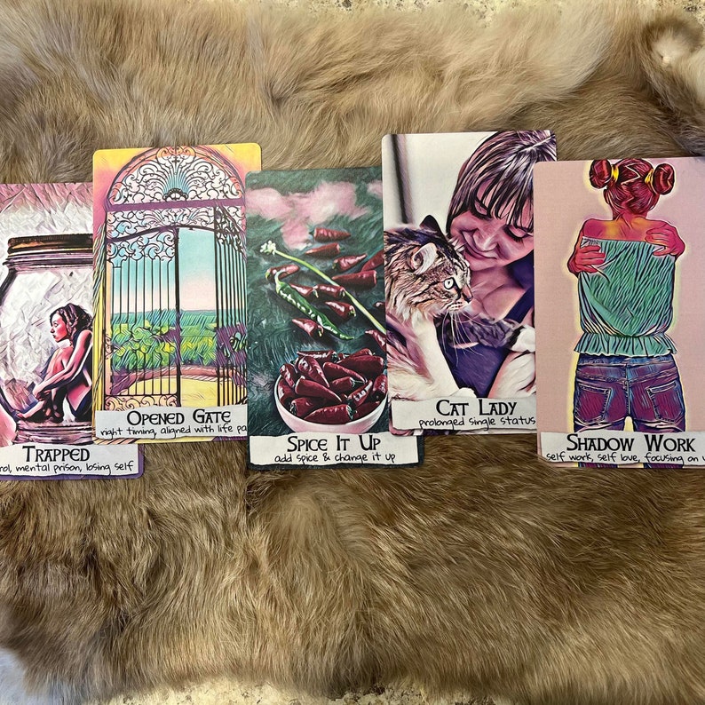 May include: Five colorful cards with different images and text. The cards are laid out on a brown furry surface. The cards are from a tarot deck and include images of a jar, a gate, red peppers, a cat and a woman, and a woman hugging herself. The text on the cards reads "Opened Gate", "Trapped", "Spice It Up", "Cat Lady", and "Shadow Work".