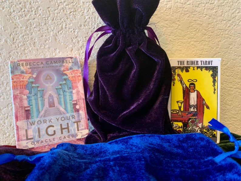May include: A purple velvet bag with a drawstring closure, a blue velvet bag, and two decks of cards. One deck is titled "Work Your Light Oracle Cards" by Rebecca Campbell. The other deck is titled "The Rider Tarot" with the number "I" on the card.