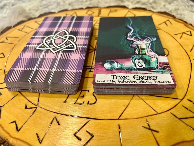 May include: Two tarot cards on a wooden surface with a circular design. The card on the left has a purple and pink plaid pattern with a white Celtic knot design. The card on the right has a green and black image of a bottle with smoke coming out of it. The text on the card reads "Toxic Energy unhealthy behavior, abuse, trauma".