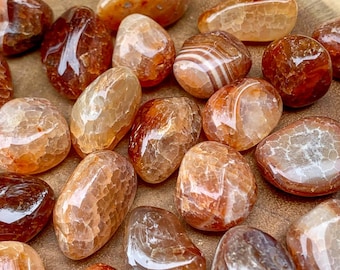 May include: A collection of carnelian tumbled stones. The stones are a mix of orange, red, and brown colors with white banding. The stones are smooth and polished.